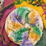 Colorful Mardi Gras King Cake topped with purple, green, and gold sugar, surrounded by festive beads and a carnival mask.