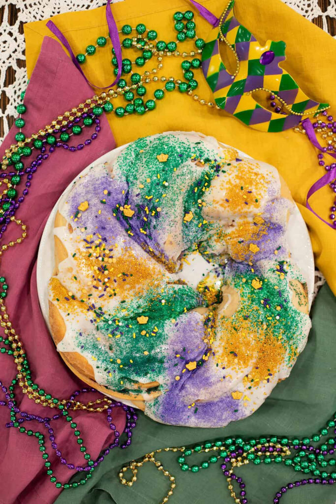 Colorful Mardi Gras King Cake topped with purple, green, and gold sugar, surrounded by festive beads and a carnival mask.