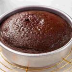 Freshly baked chocolate cake in a round silver cake pan, resting on a cooling rack